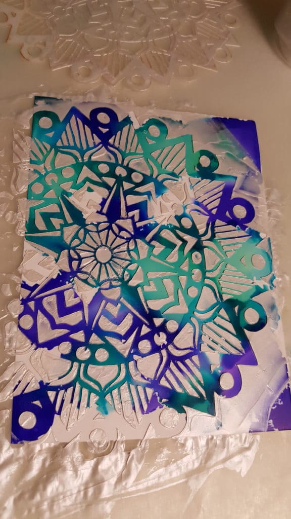 Yupo paper with blue, turquoise, and purple alcohol inks that has been stencilied over using shimmery goodness applied with a palette knife through the striped mandala stencil.