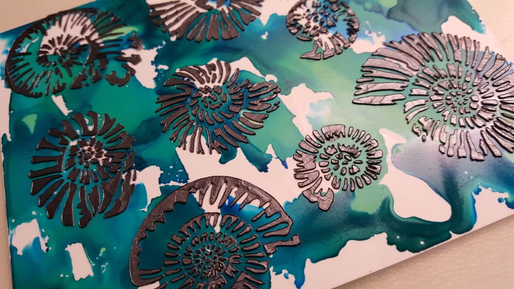 Marcasite silver modeling paste applied with a palette knife through the TCW500 Nautilus stencil on top of blue and green alcohol inks on yupo paper.