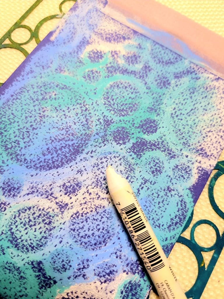 Close up image showing the stencil beneath the paper. A white NeoColour II crayon is laying on top of the textured paper which has had the crayons applied to it.